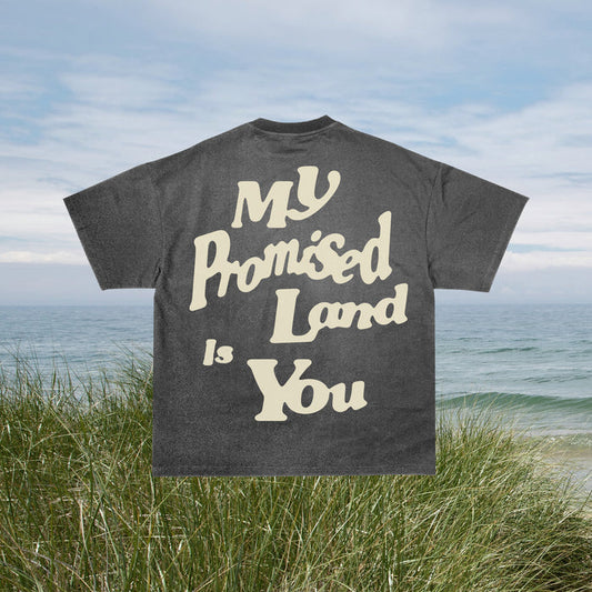 A gray t-shirt with the text 'MY PROMISED LAND IS YOU' printed on the front, displayed on a hanger against a beach backdrop.